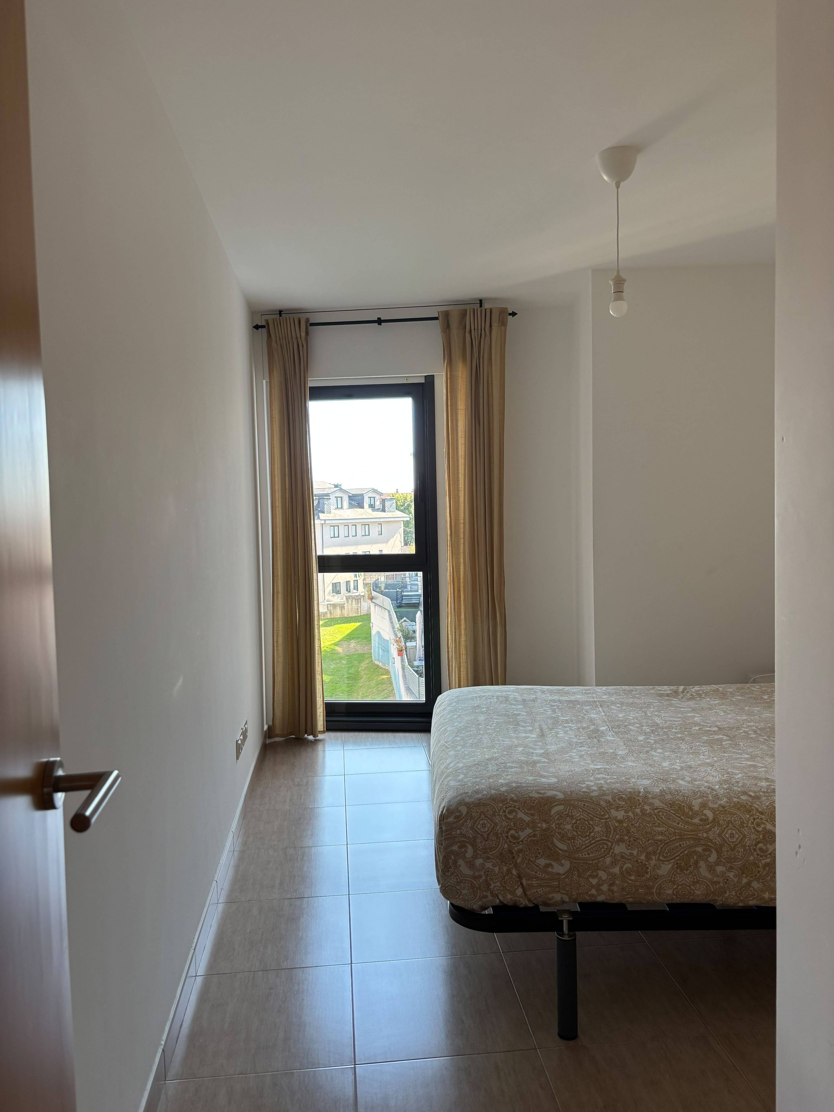Un dormitorio con cama, una puerta, un ventanal con cortinas y vistas a edificios.