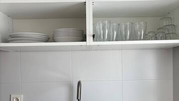 Una cocina con un lavamanos de acero inoxidable, un tapete para escurrir trastes y estantes con platos, tazones y tazas.