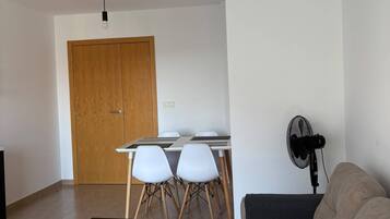 Una sala de estar con un sofá, una mesa de centro, una mesa de comedor con sillas, una puerta de madera y un ventilador de pie.