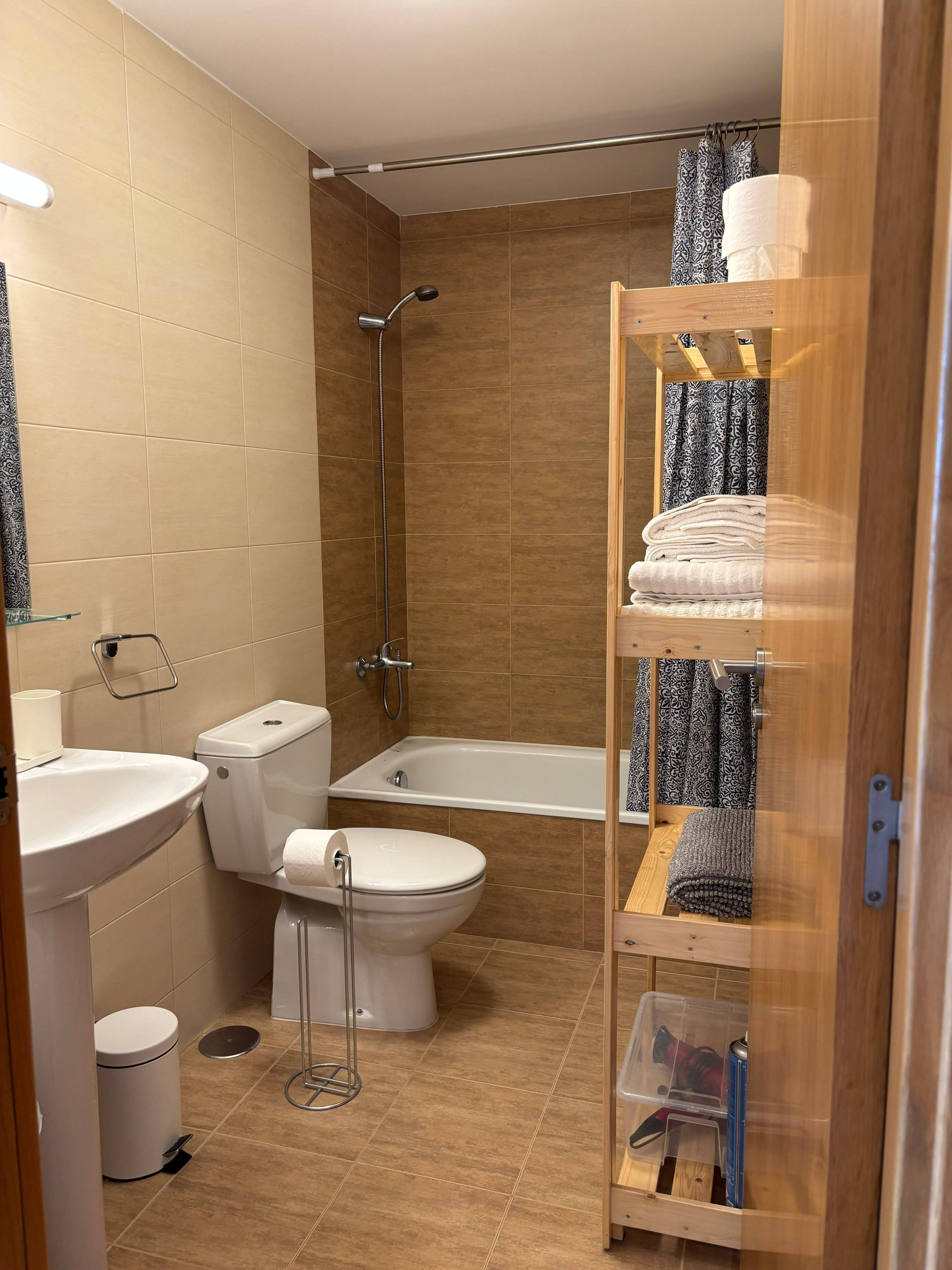 Baño con inodoro, lavabo, bañera y ducha. Hay un perchero con toallas y un estante con artículos de aseo.