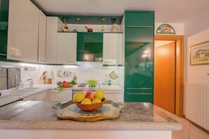 Een keuken met witte en groene kastjes, een fruitschaal op het aanrecht en een deur naar een andere kamer.