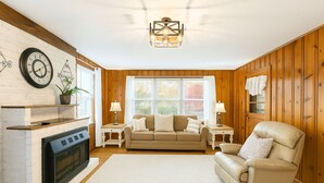 A cozy living room with a fireplace, wooden walls, a sofa, a recliner, and a chandelier. - Spacious Family Retreat  2 Indoor Fireplaces + Volleyball Fun! (Flat Rock)