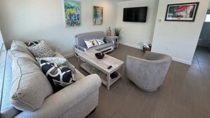 A modern living room with a grey sofa, a coffee table, and a grey armchair. There is a flat-screen TV mounted on the wall, and a painting above the sofa. - "St.Thomas"-Family Villa-2 Blks 2 Beach-BeachGear-2 BR 2 BA-Tranquil Courtyard (Bradenton Beach)