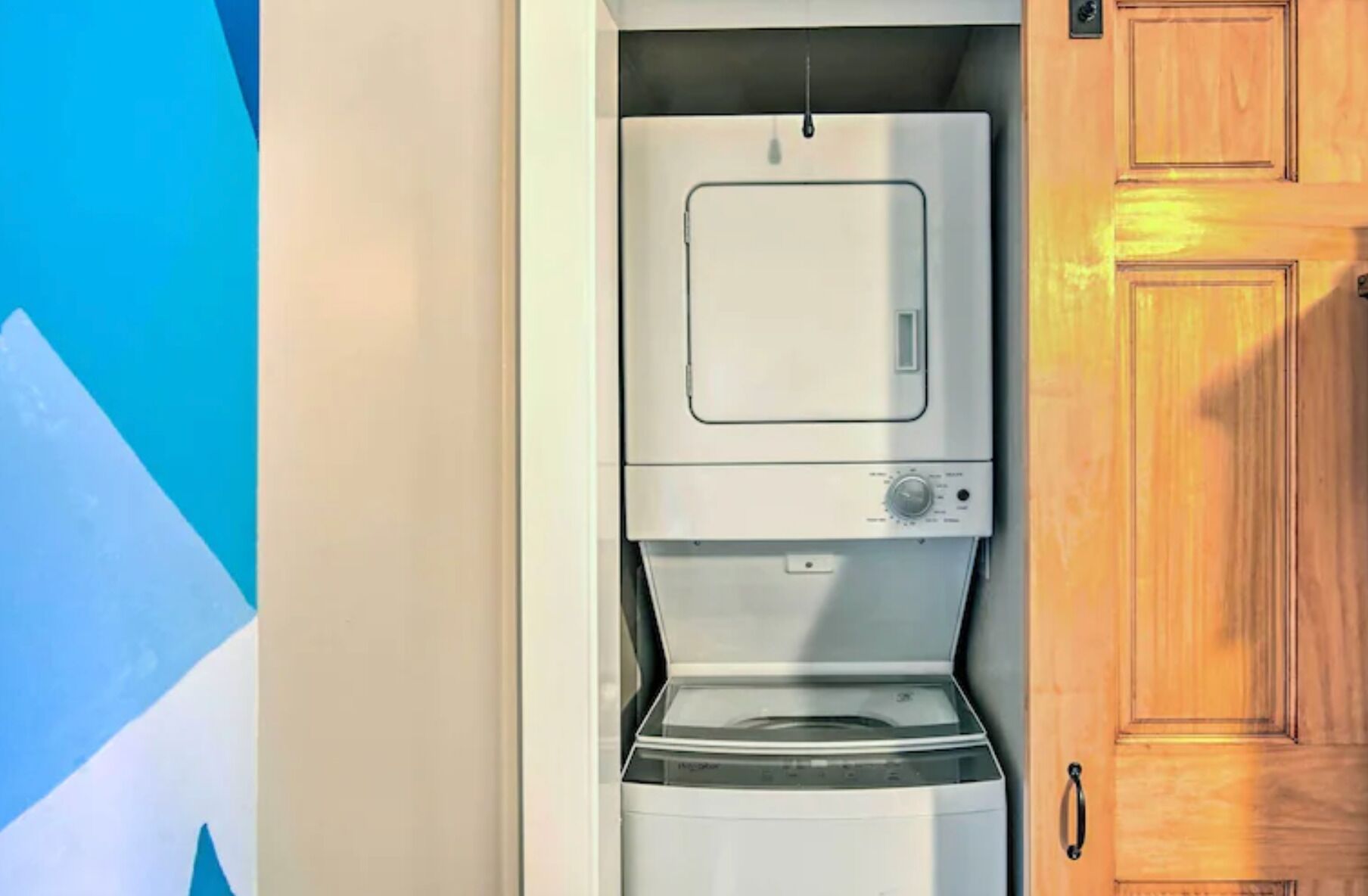Un appareil compact combinant lave-linge et sèche-linge, installé dans une pièce dotée d’une porte en bois et d’un mur bleu.