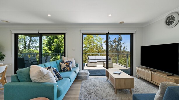 A modern living room with a sofa, coffee table, and TV, featuring large sliding glass doors to a balcony with outdoor furniture. - Fern & Maple Lodge Wentworth Falls (Wentworth Falls)
