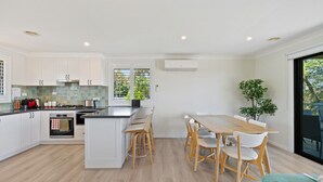 A modern kitchen with white cabinets, a central island, and a dining area with wooden chairs. - Fern & Maple Lodge Wentworth Falls (Wentworth Falls)