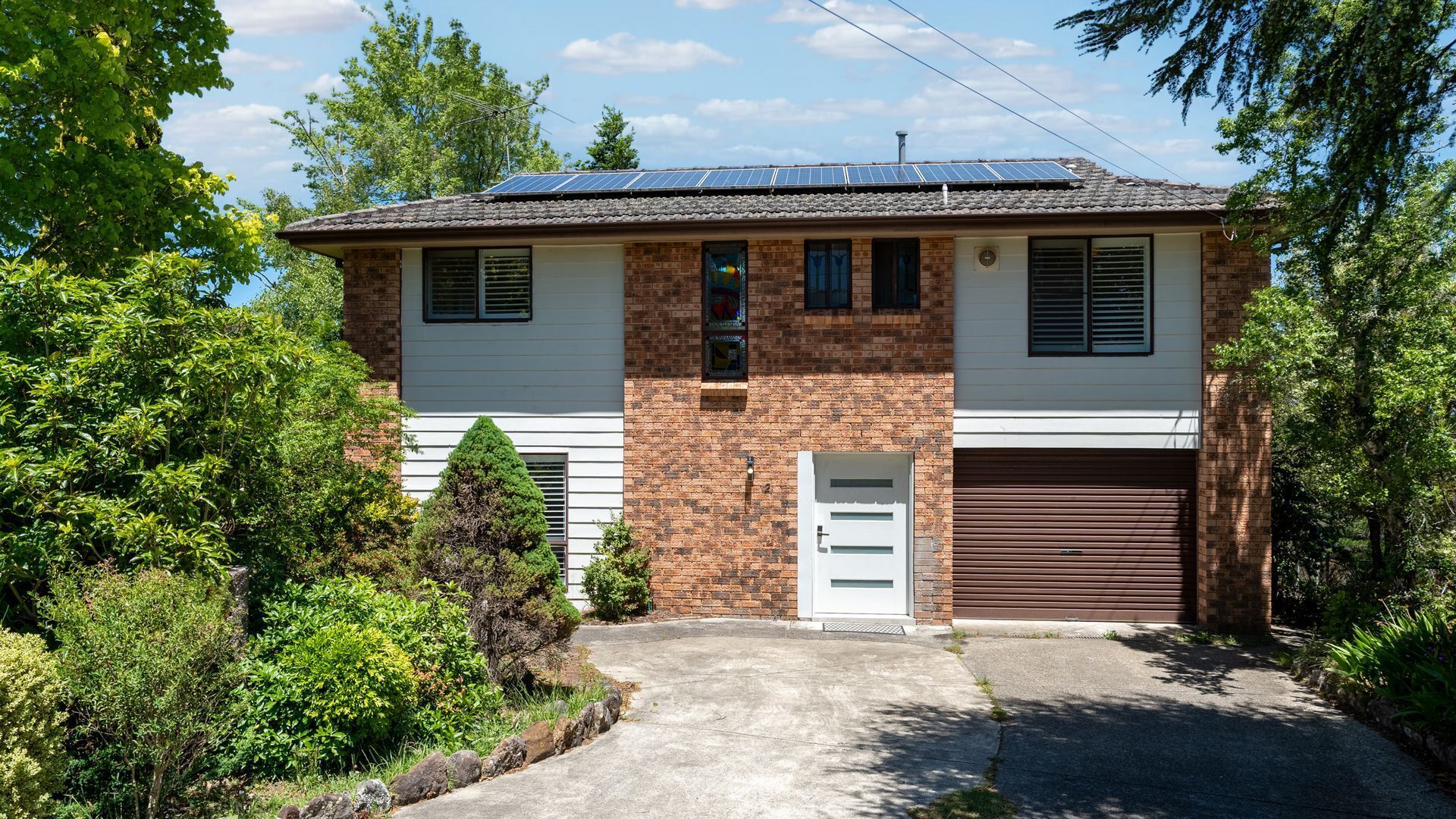 Une maison à deux étages avec une façade en briques, des panneaux solaires sur le toit, un garage et une allée menant à l’entrée.