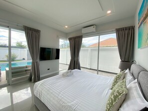 A bedroom with a large bed, a flat-screen TV, a ceiling fan, and a view of a swimming pool. - Modern Villa 3BR - Great Location KL3 (Hua Hin)