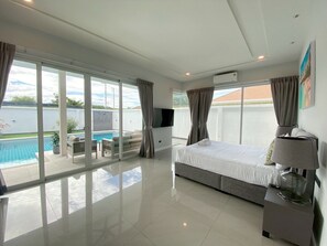 A modern hotel room with a large bed, a TV, a bedside table with a lamp, and a view of a swimming pool and outdoor seating area. - Modern Villa 3BR - Great Location KL3 (Hua Hin)