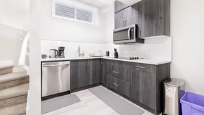 A compact kitchen with dark wood cabinets, a stainless steel dishwasher, a microwave, and a coffee maker. - Charming 2Bed Retreat near Airport & Banff w/ desk (Airdrie)
