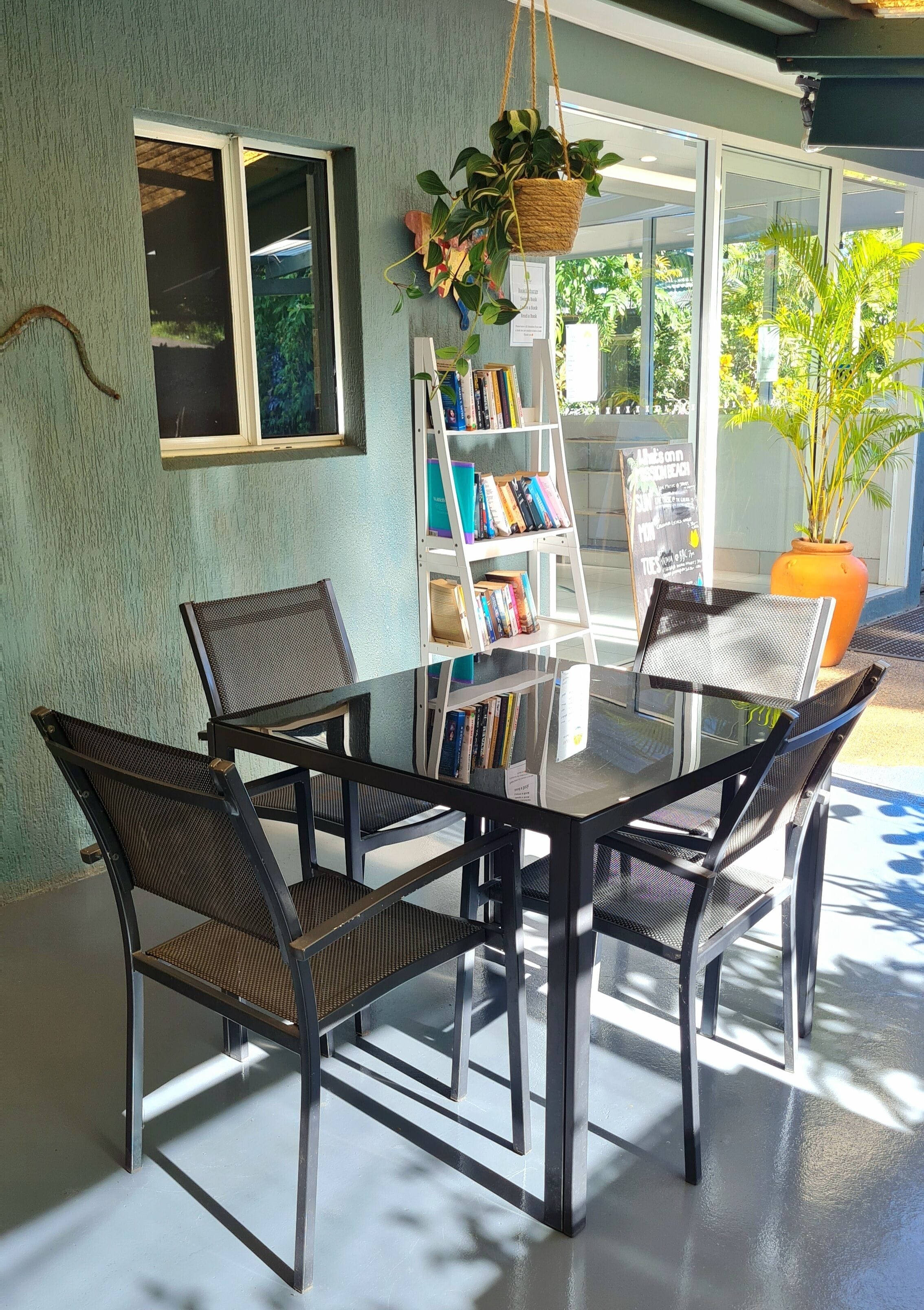 A glass-top table with four chairs, a bookshelf, and a hanging plant in a well-lit room with a view of greenery outside.