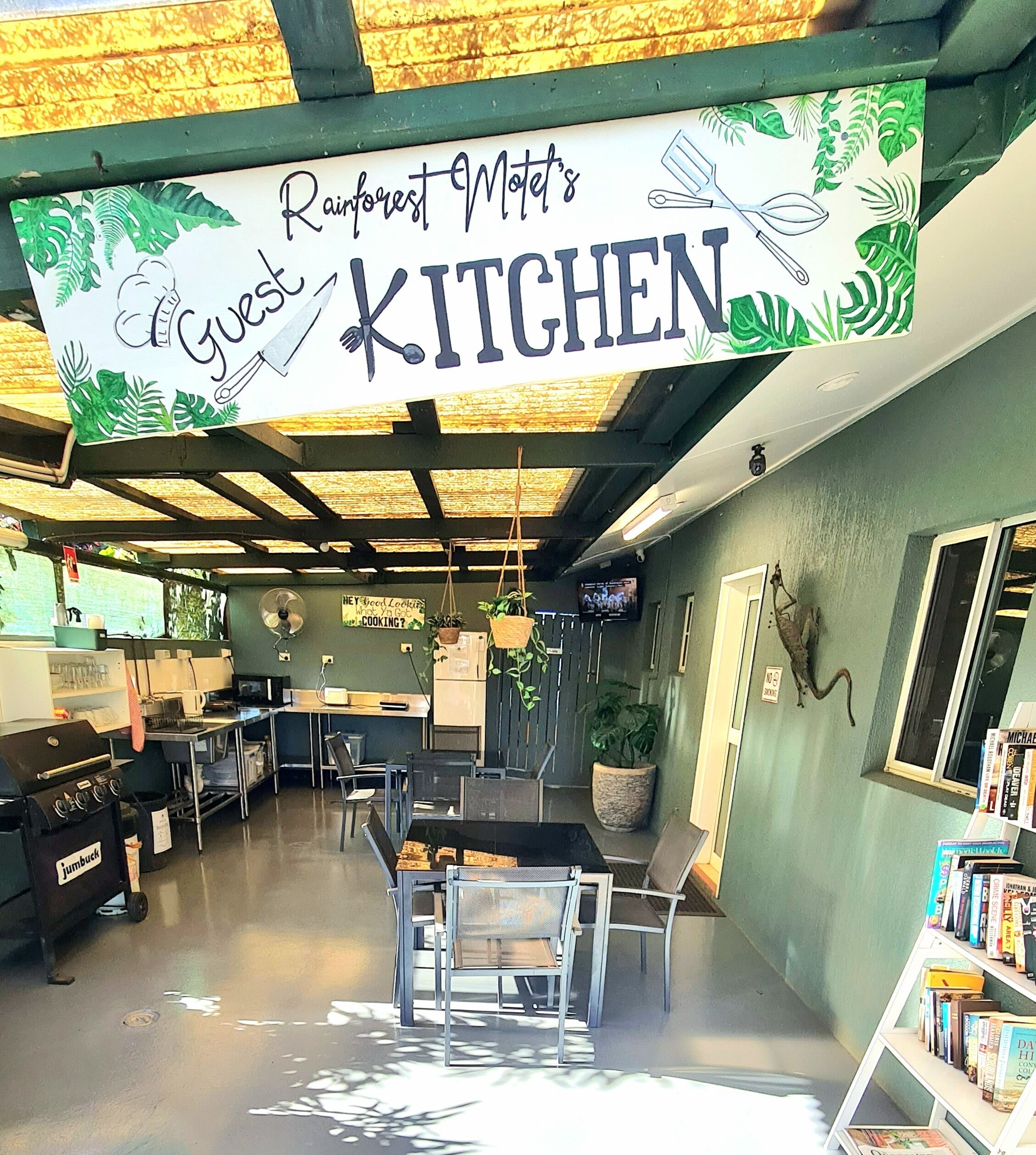 Et tekøkken med et skilt der står "Rainforest Motel's Guest Kitchen", med et rustikt loft, en kaffemaskine, en vask og en reol med bøger.