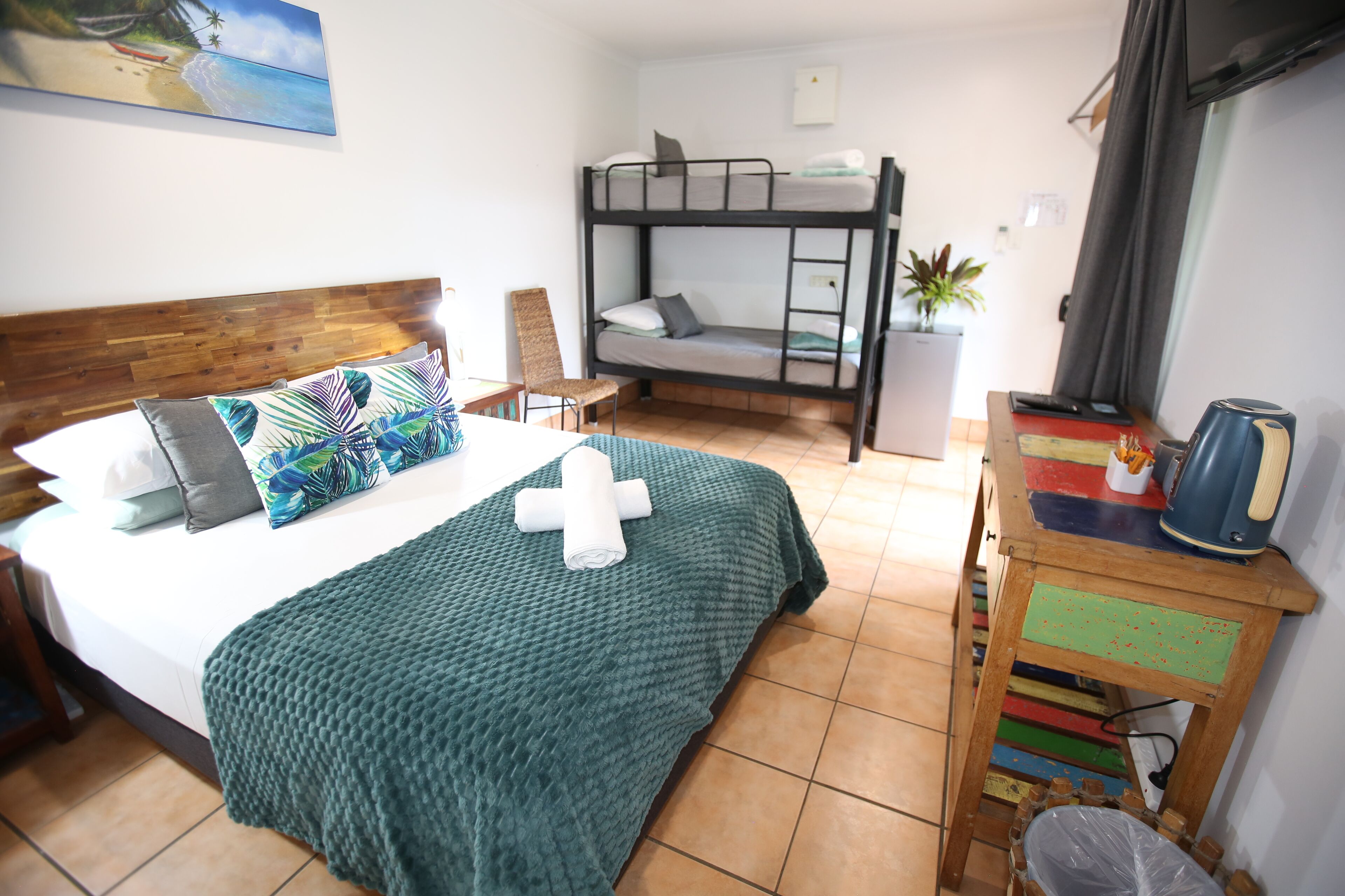 A bedroom with a bunk bed, a single bed with a green blanket, a wooden headboard, a chair, a small table with a kettle, and a painting of a beach.