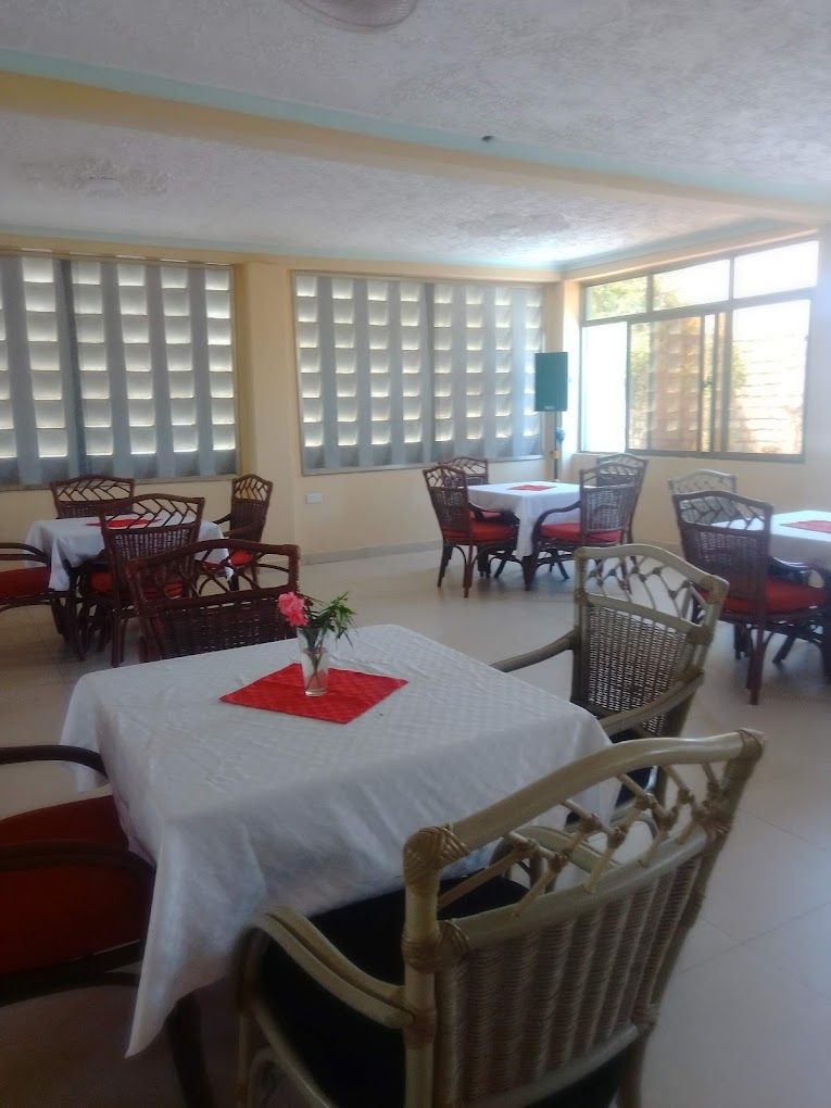 A dining area with tables and wicker chairs, large windows with blinds, and a ceiling fan.