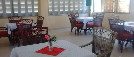 A dining area with tables and wicker chairs, large windows with blinds, and a ceiling fan.