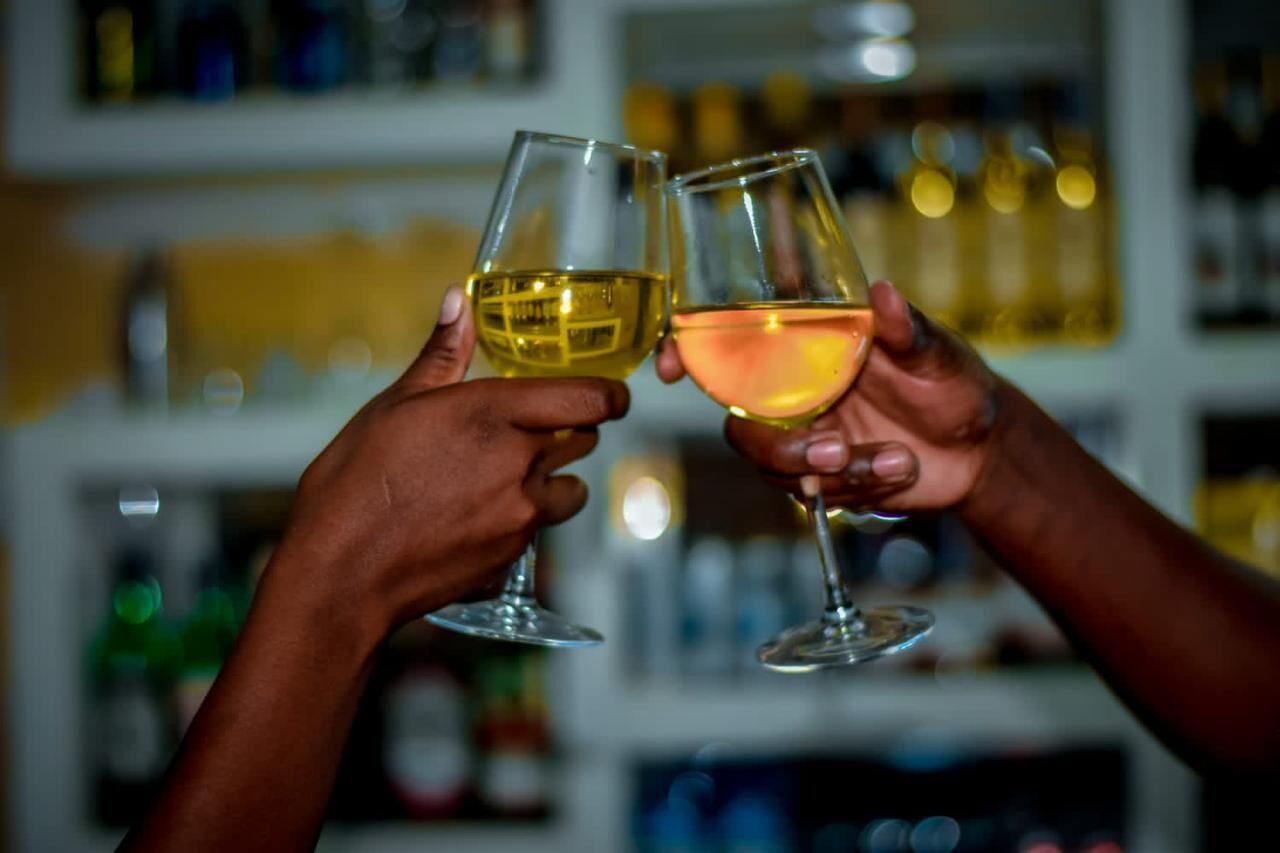 Two hands holding wine glasses, one with a yellow liquid and the other with a pinkish liquid, in a bar setting with shelves in the background.