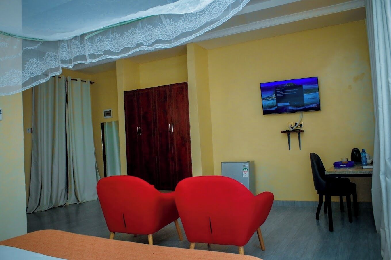 Habitación de hotel con una cama, dos sillas rojas, un televisor en la pared, un escritorio con una silla y un armario.
