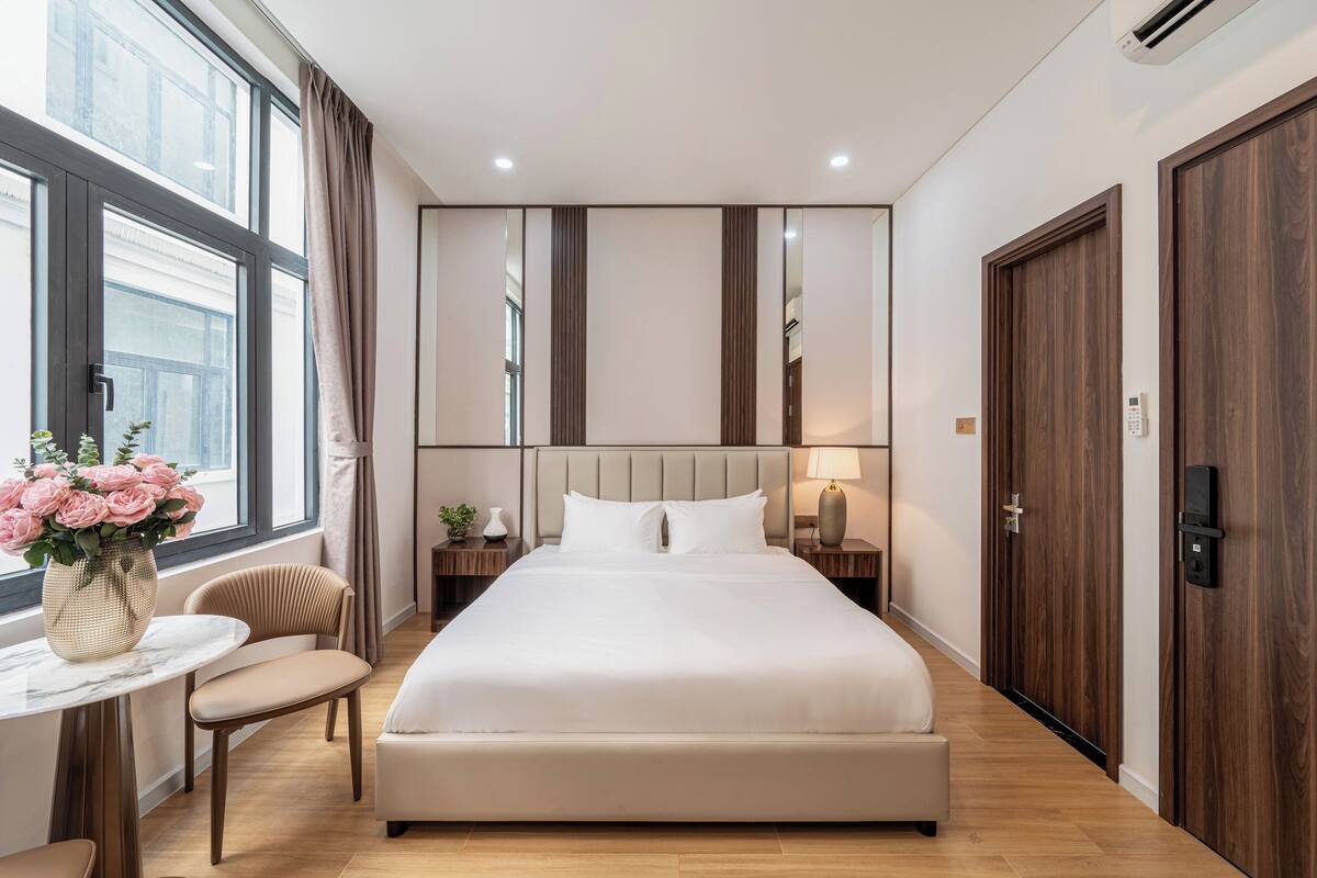 A modern hotel room with a large bed, a bedside table with a lamp, a chair, and a window with curtains.