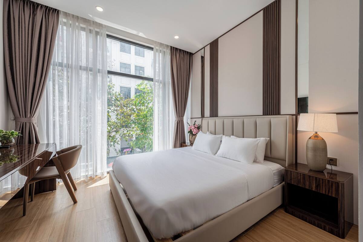 A modern hotel room with a large bed, a desk with a chair, a nightstand with a lamp, and a view of greenery outside.