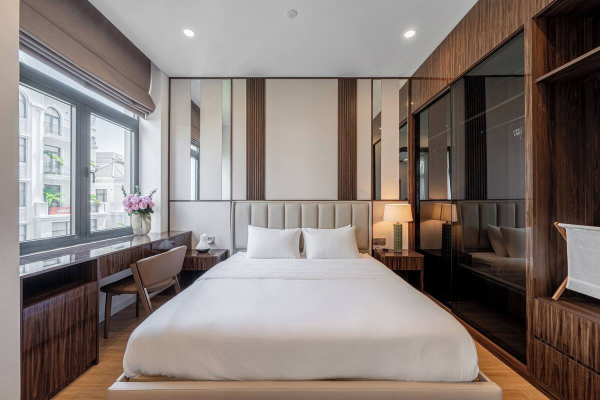A modern hotel room with a large bed, a desk, and a view of the city through large windows.