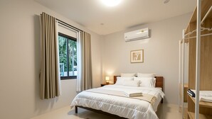 A bedroom with a bed, a bedside table, an air conditioning unit, a window with curtains, and a wooden wardrobe. - Nirvana Phangan Villas (Tambon Ko Pha-ngan)