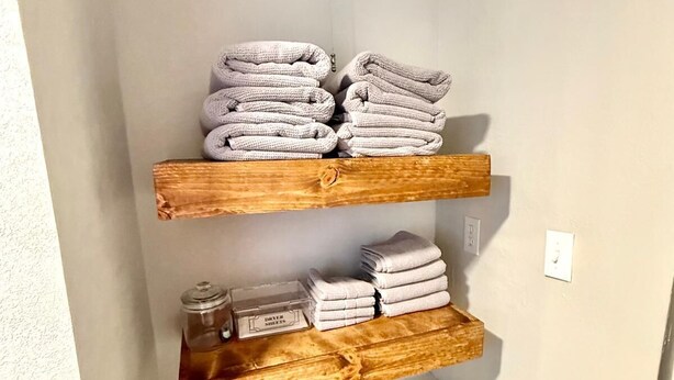 Een badkamer met houten planken waarop opgevouwen handdoeken en een bakje met de tekst 'DRYER SHEETS' staan.