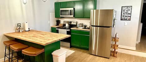 Cozinha moderna com armários verdes, geladeira de aço inoxidável, ilha de madeira com banquetas e iluminação pendente.