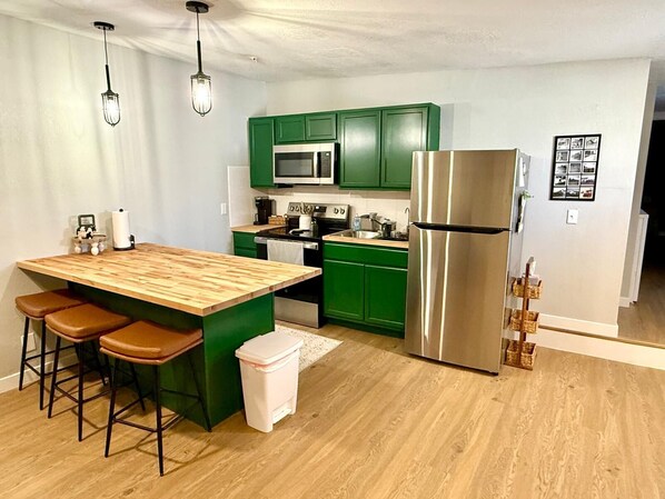 A modern kitchen with green cabinets, a stainless steel refrigerator, a wooden island with stools, and pendant lighting.