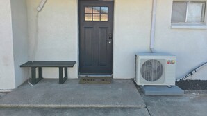 Een huisdeur met een donkere deur, een vloermat, een bankje en een airconditioning.