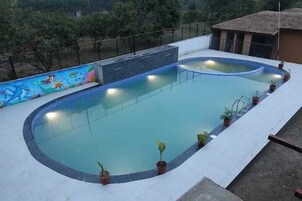 Piscina com um mural, rodeada por plantas em vasos, e uma estrutura de telhado de palha ao fundo.