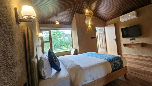 Une chambre avec un plafond en bois, un lit recouvert de draps blancs, un plaid bleu, un téléviseur fixé au mur et une fenêtre donnant sur un paysage verdoyant.