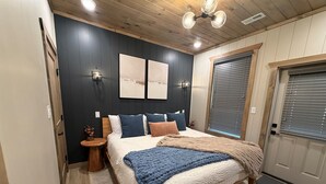 Une chambre à coucher avec un lit en bois, une table de chevet, une lampe fixée au mur et une fenêtre avec des stores.