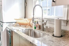 A kitchen with a stainless steel sink, a wooden drying rack, and a window with a view of buildings. - Sojourn | Coastal Cottage | 3 BR (Norfolk)