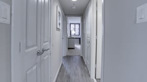 A narrow hallway with white doors and a wooden floor, leading to a kitchen area with a window. - Sojourn | Courtland Towers w/ racquetball courts (Arlington)