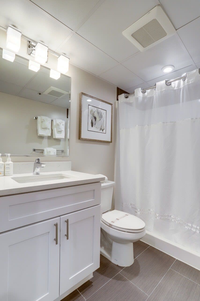 A modern bathroom with a white sink, mirror, and toilet. There is a shower curtain and a framed picture on the wall.