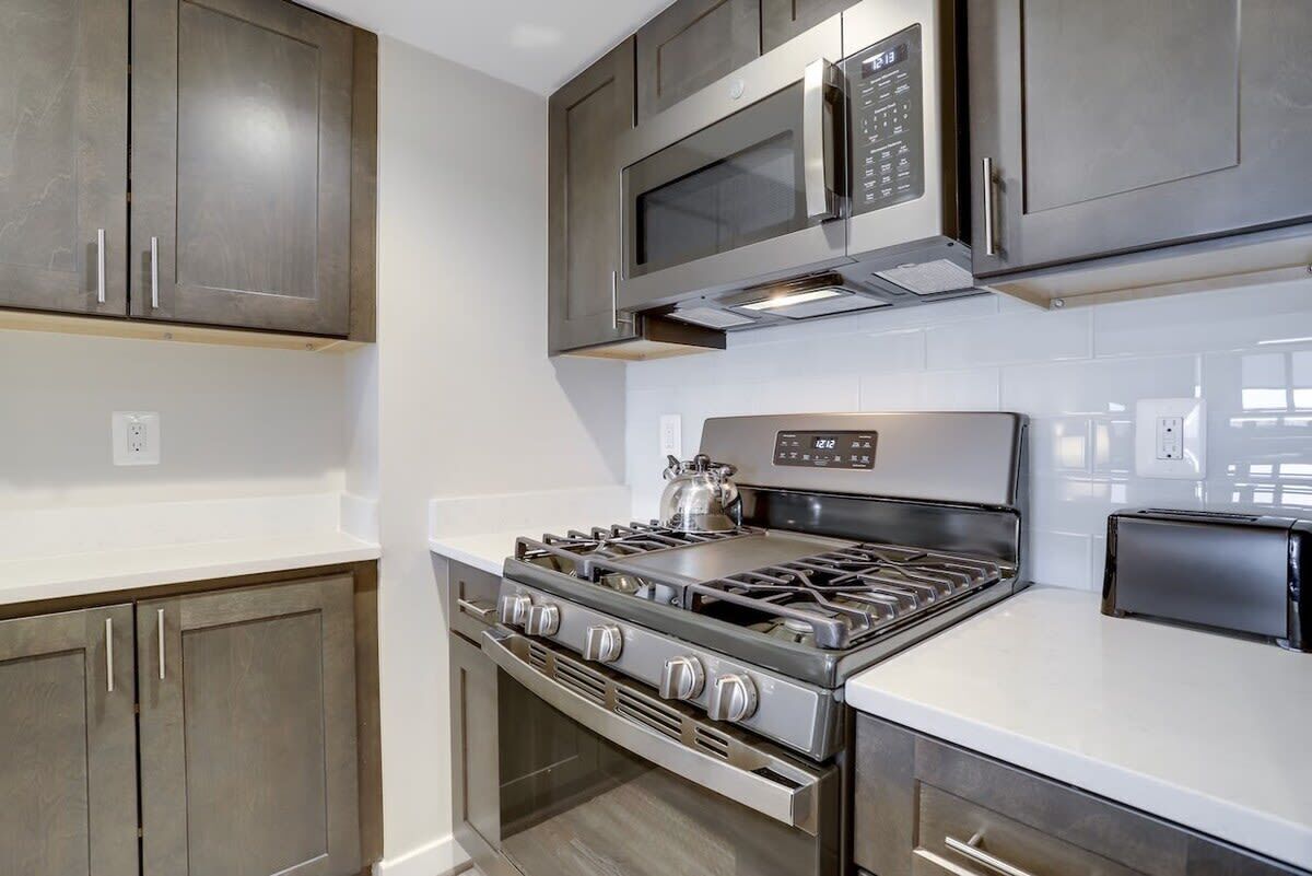 A modern kitchen with stainless steel appliances, including a stove, microwave, and oven, with wooden cabinets and white countertops.