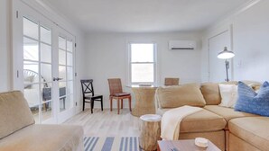 Uma sala de estar ampla com um sofá bege, uma mesa de centro de madeira e um tapete listrado. Há uma área de jantar com uma mesa redonda e cadeiras, e uma grande janela que oferece vista para o exterior.