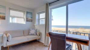 Quarto com sofá branco, escrivaninha de madeira e uma grande janela com vista para a praia.