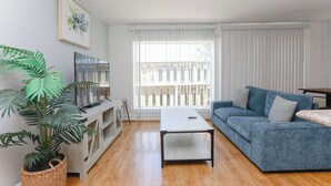 A living room with a blue sofa, a white coffee table, a TV on a stand, a large window with blinds, and a potted plant. - Sojourn Willoughby Spit Chic Shack <1min. to Beach (Norfolk)