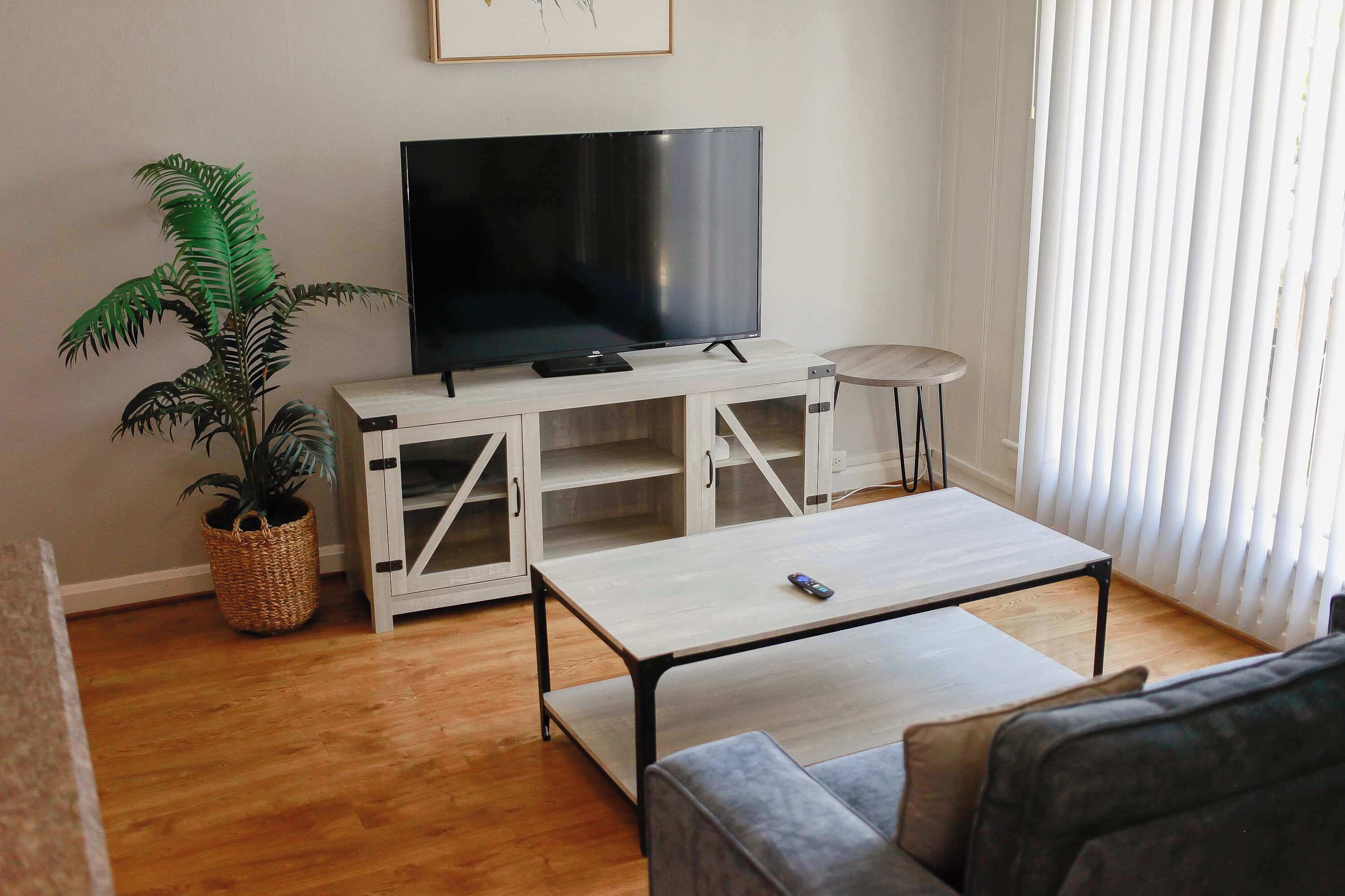 Un soggiorno con TV a schermo piatto su un mobile bianco, un tavolino, un divano, una pianta in vaso e una finestra con persiane.