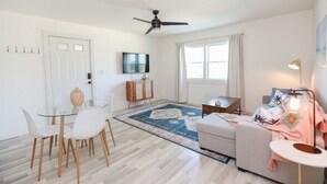 Una sala de estar moderna con una mesa de comedor de vidrio, sillas blancas, un sofá, un televisor y un ventilador de techo.