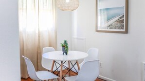 Et spiseområde med et rundt bord, hvide stole, en vævet pendel, et indrammet billede på væggen og et vindue med lette gardiner.