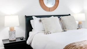 En pænt redt seng med hvide sengetøj og to puder, placeret mellem to sengeborde med lamper, hvoraf det ene har et geometrisk mønster.