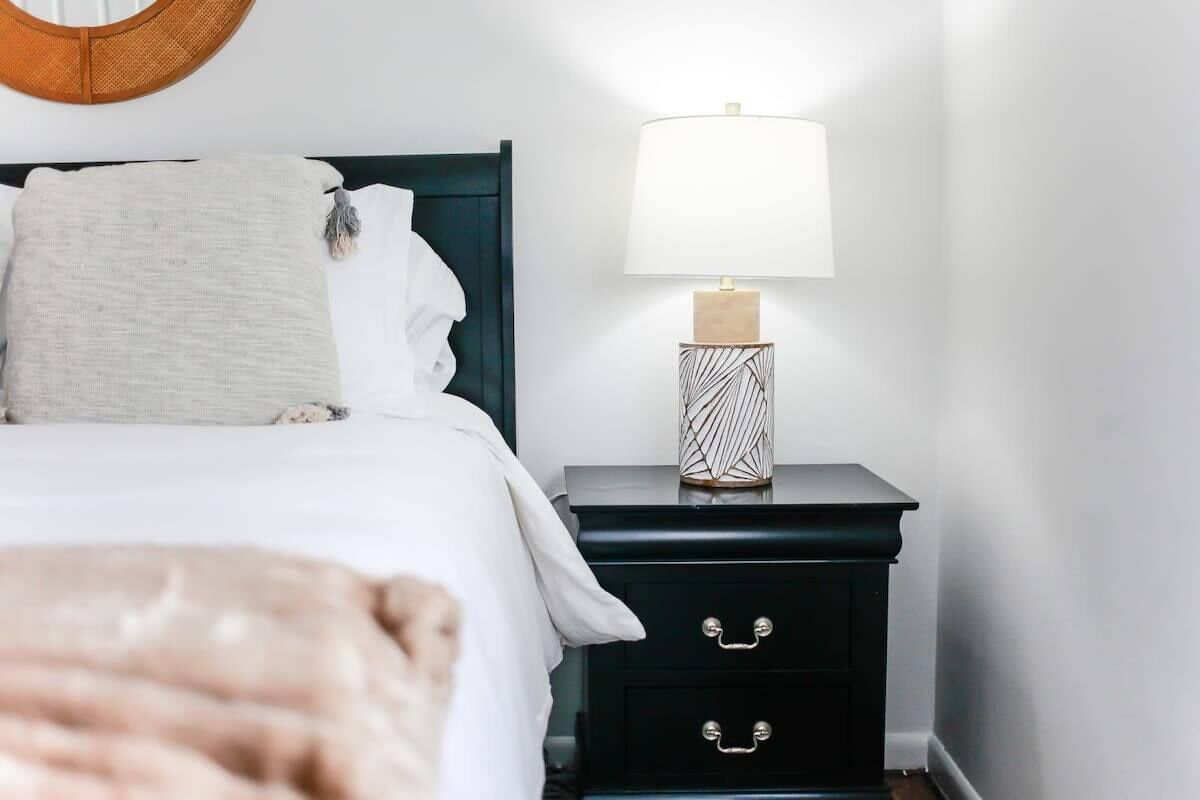 A neatly made bed with white linens and a beige throw blanket, a bedside table with a lamp and a decorative vase, and a dark-colored nightstand with two drawers.