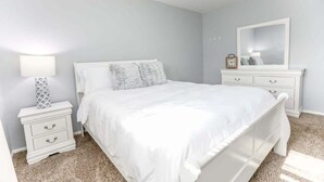 Ein Schlafzimmer mit einem weißen Bett, einem Nachttisch mit Schubladen, einer Kommode mit Spiegel und einer Lampe.