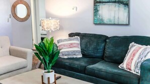 Ein Wohnzimmer mit einem dunkelgrünen Sofa, einem weißen Sessel, einem runden Spiegel, einer Lampe und einem Bild an der Wand.