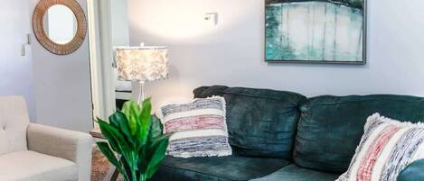 Una sala de estar con un sofá verde oscuro, un sillón blanco, un espejo redondo, una lámpara y un cuadro en la pared.