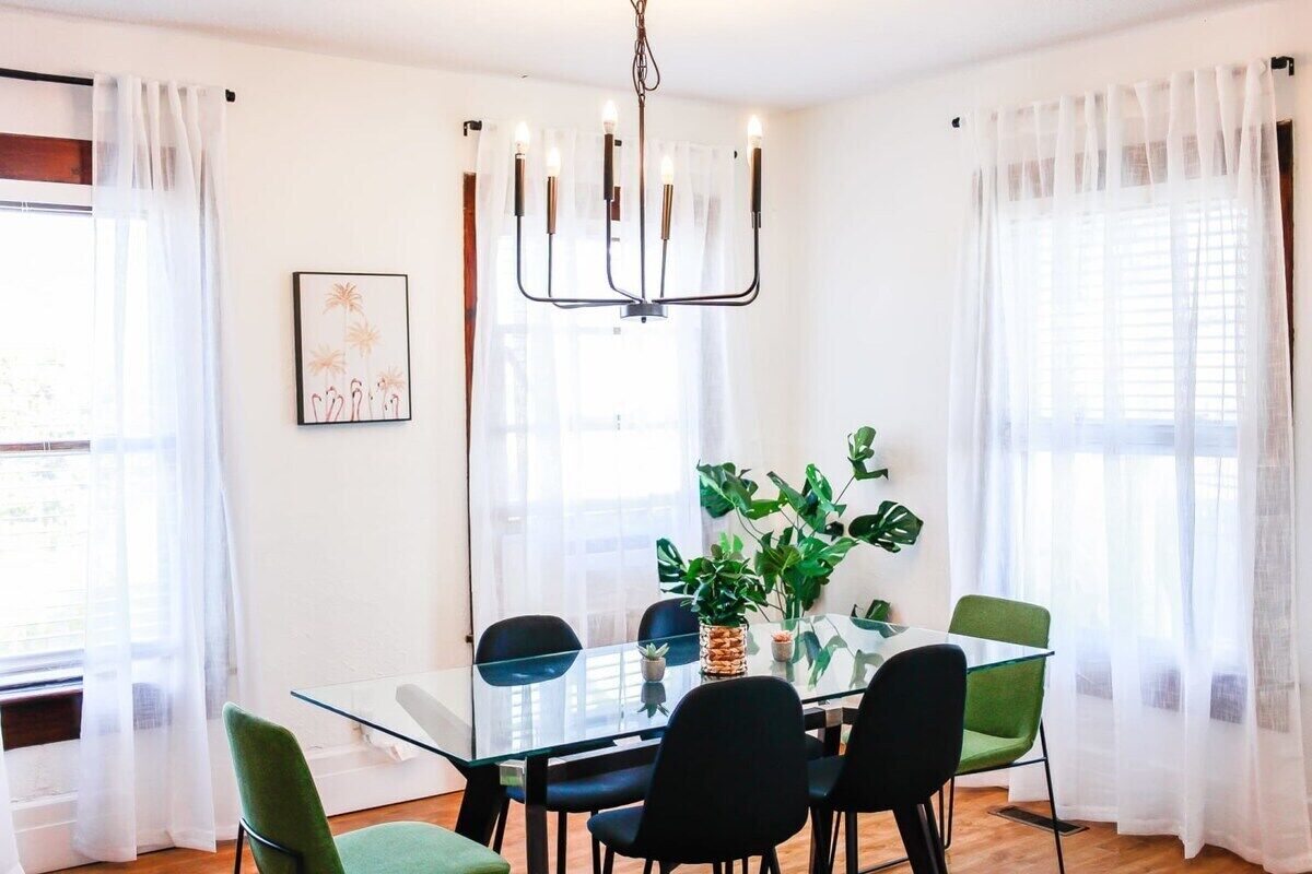 A dining area with a glass table, black chairs, and a green chair. There is a chandelier above the table and a plant on the table.