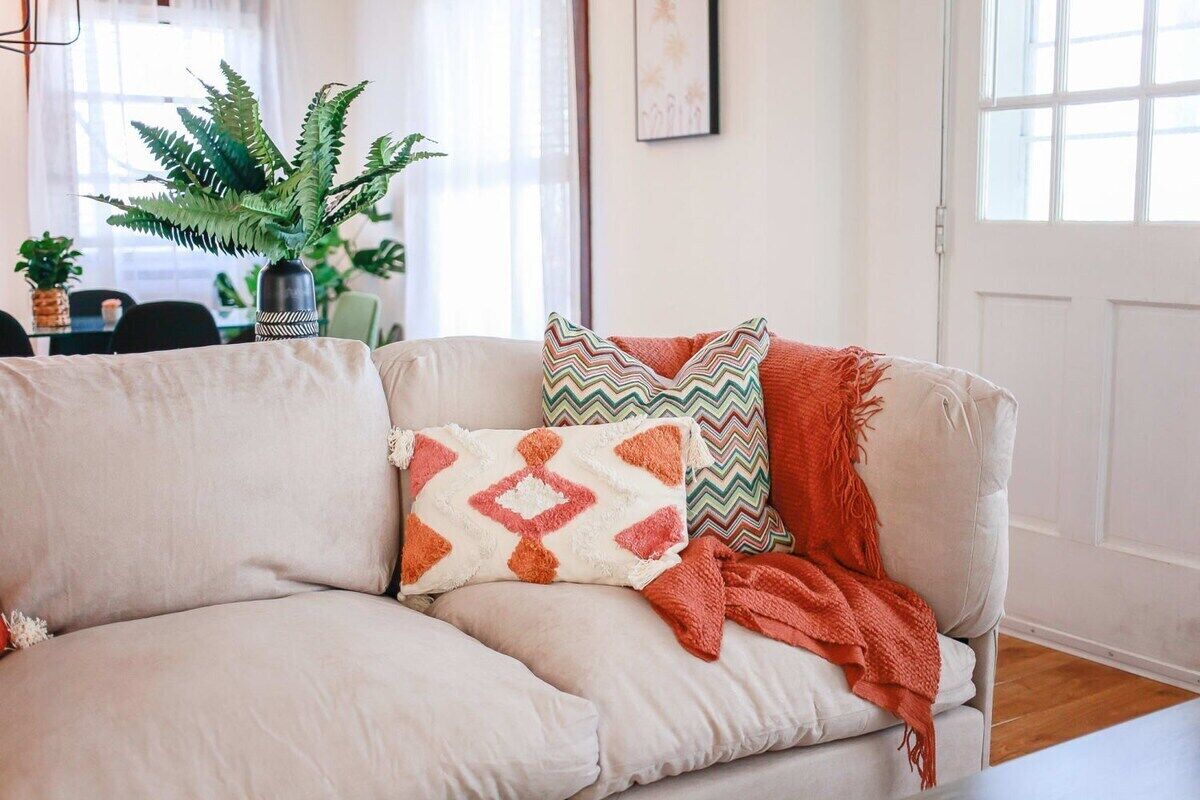 Un canapé beige agrémenté de coussins à motifs et recouvert d’un plaid, une plante en pot et un tableau encadré accroché au mur.