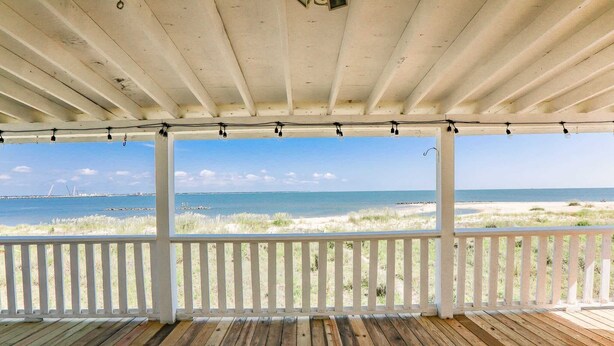 Une terrasse en bois avec une balustrade blanche, donnant sur une plage bordée de dunes et l’océan.